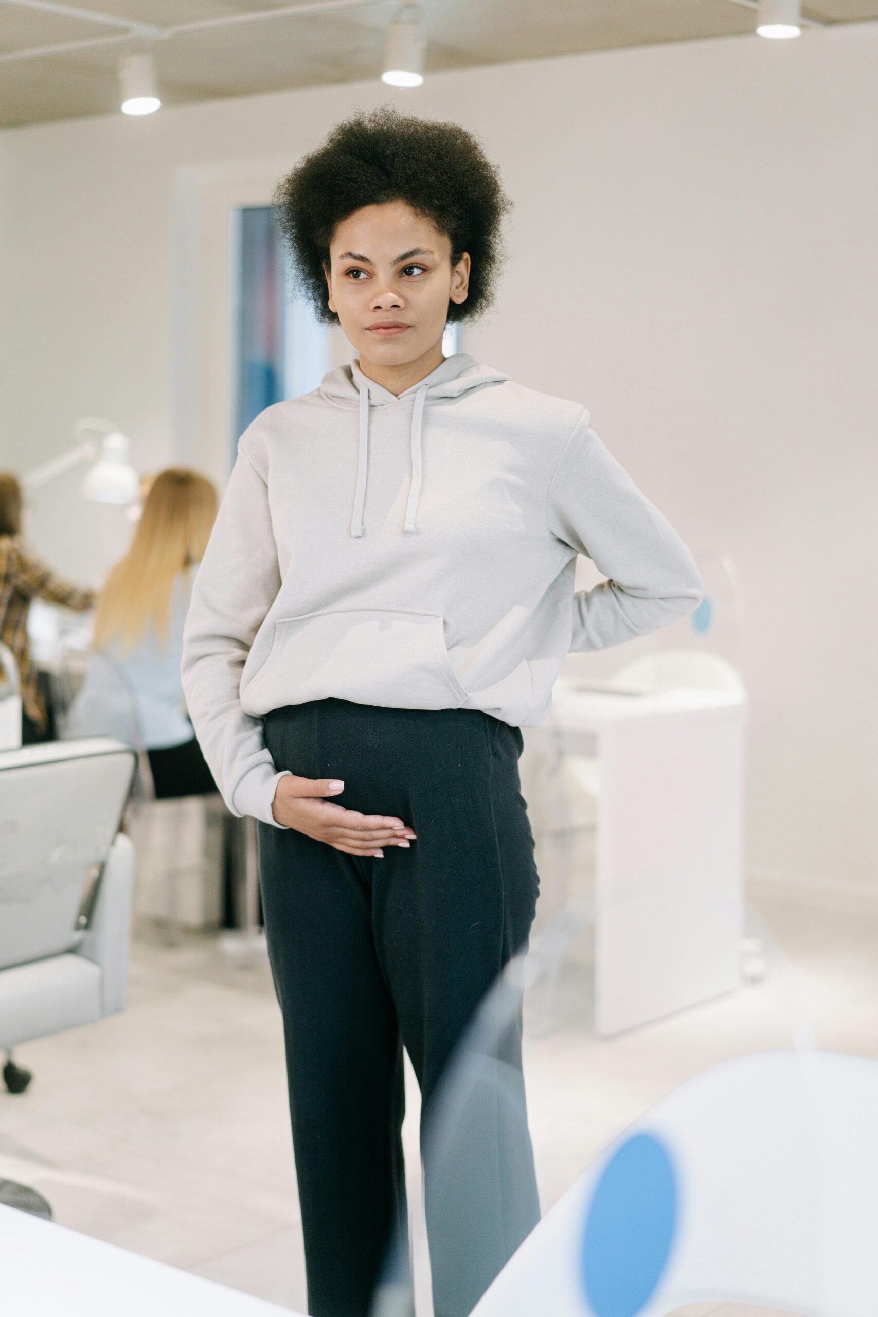 Pregnant woman standing in an office, hand on belly, wearing a hoodie. Modern workspace environment.