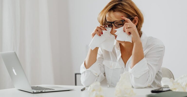 A woman in glasses is wiping tears while seated at a desk with a laptop and tissues.