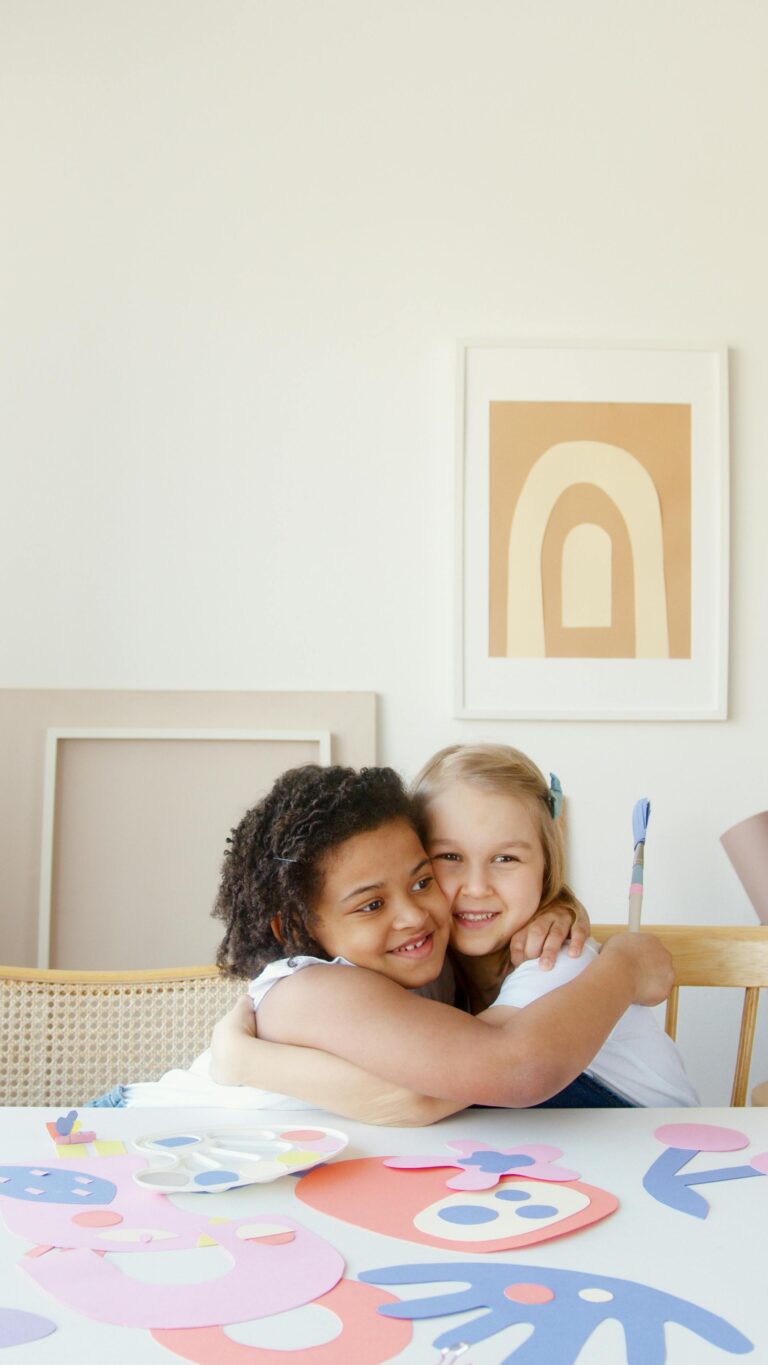 Two happy children hugging in a bright art classroom with colorful papers.